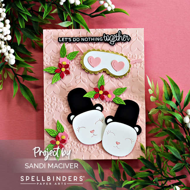 Handmade card featuring two adorable panda bear slippers nestled together on a blush pink background embossed with the Sweet Stems Embossing Folder from the Spellbinders December Club kits. Die-cut flowers and leaves decorate the card, with a coordinating sleep mask above the slippers and a black-and-white sentiment that reads “Let’s do nothing together.” Card created using the Spellbinders Deluxe Caboodle Bonus Gift of the Month.