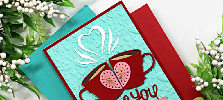 Handmade Valentine’s card featuring two red stitched coffee mugs with pink heart centers created using the Spellbinders Stitched Die of the Month “A Perfect Blend.” The mugs meet in the middle with white steam forming a heart above them. The sentiment “love you” is die-cut in red with a pink banner reading “Let’s Chat Soon.” The aqua card front is embossed with the Unlock My Heart 3D Embossing Folder and layered on red and aqua cardstock, surrounded by greenery and white berry accents.