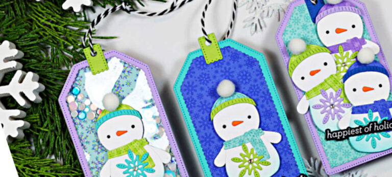 Three handmade die-cut snowman holiday gift tags in pastel blue, purple, and teal, featuring layered snowmen, snowflake accents, twine ties, and one shaker tag, styled on greenery with white snowflake decorations.