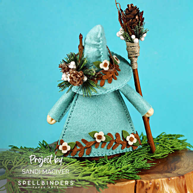 Back view of a handmade felt gnome in a teal coat and hat, decorated with pinecones, greenery, and stitched leaf and flower embellishments, standing on evergreen branches with a rustic wood staff.
