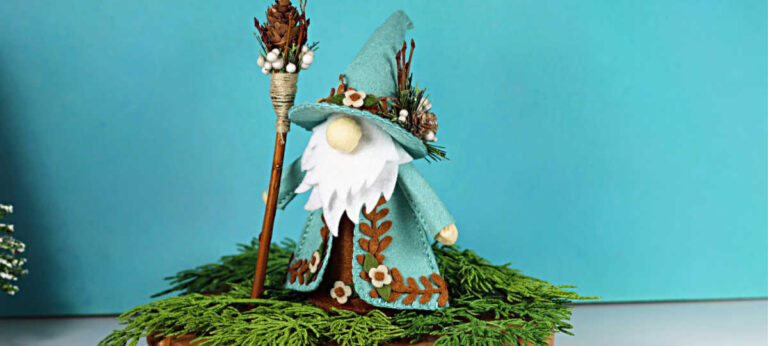 Handmade felt gnome standing on greenery with a pinecone staff, teal coat, and stitched leaf embellishments; Woodlands Fergus project created using Spellbinders dies.