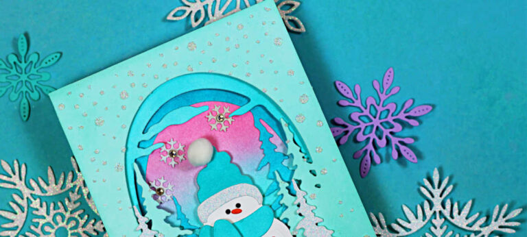 Handmade winter birthday card featuring an easy diorama design with a smiling snowman wearing a teal scarf and hat, standing inside a layered arched window. The scene includes snowy pine trees, falling snow accents, and a pink-to-teal blended sky, set on a teal card base with scattered white splatter and a small “Happy Birthday” sentiment.