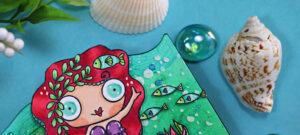 Under-the-sea themed MemoryDex card created with AALL & Create stamps and dies, featuring a whimsical red-haired mermaid with teal scales seesawing above a row of small fish. The mixed-media background is ink-blended in aqua and turquoise tones with splatters, sea plants, coral, and shells, accented with sequins and a “Sea Life” label.