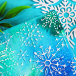 A handmade winter birthday card with a teal, blue, and green watercolor-style background created using Lindy’s Magical Powders and the Simon Says Stamp Wonderland Snowflakes embossing folder. White snowflake designs are embossed across the panel, and a layered “Happy Birthday” sentiment in white cardstock and vellum sits in the center, giving the card a soft, snowy, elegant look.
