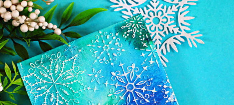 A handmade winter birthday card with a teal, blue, and green watercolor-style background created using Lindy’s Magical Powders and the Simon Says Stamp Wonderland Snowflakes embossing folder. White snowflake designs are embossed across the panel, and a layered “Happy Birthday” sentiment in white cardstock and vellum sits in the center, giving the card a soft, snowy, elegant look.