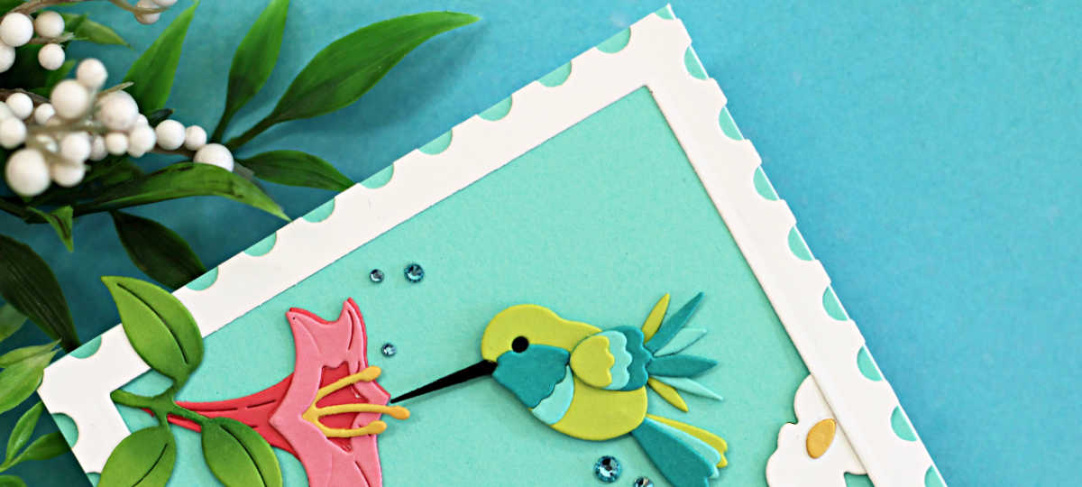 Die-cut handmade card featuring a colorful hummingbird hovering beside a pink flower over white daisies and layered green grass, framed with a postage-style border on a teal background with a simple “hello” sentiment at the bottom.