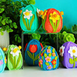 A collection of colorful handmade 3D felt Easter eggs decorated with layered felt flowers and leaves, displayed on clear risers alongside a whimsical felt gnome, set against a bright turquoise background with green foliage.
