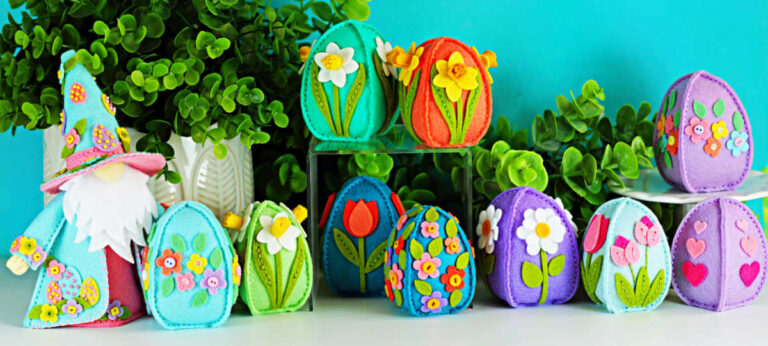 A collection of colorful handmade 3D felt Easter eggs decorated with layered felt flowers and leaves, displayed on clear risers alongside a whimsical felt gnome, set against a bright turquoise background with green foliage.