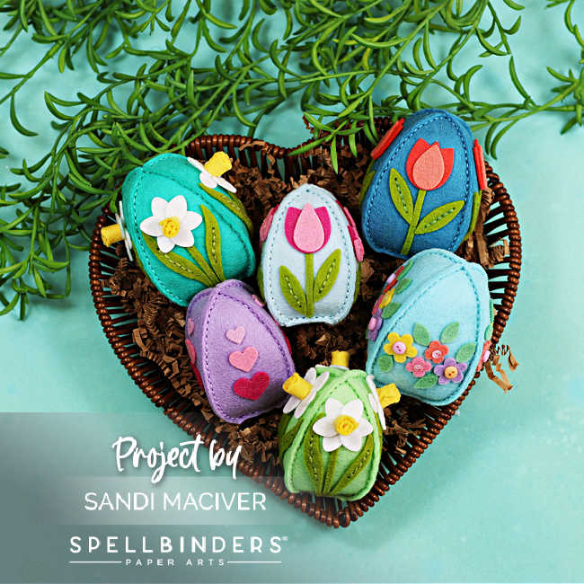 Handmade 3D felt Easter eggs decorated with flowers, hearts, and layered embellishments, arranged in a heart-shaped wicker basket with greenery on a soft aqua background.