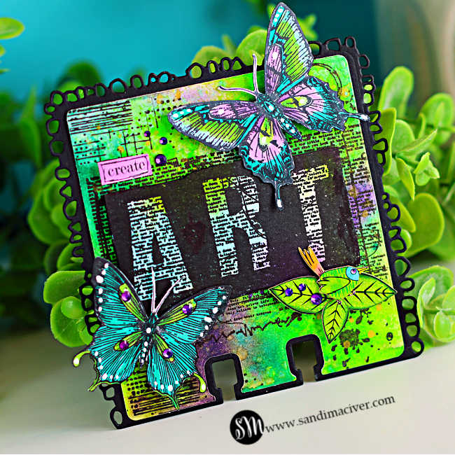Colorful mixed media display featuring a vibrant AALL & Create MemoryDex card on the right and a decorated storage box on the left. The MemoryDex card has a neon green and purple Distress Oxide spray background with bold black “ART” lettering, hand-colored butterflies in teal and purple, and a small green bird with purple rhinestone accents. Beside it, a whimsical teal box is filled with bright florals and features a red-haired mermaid illustration, playful characters, and layered collage elements. The scene is styled with lush green foliage against a teal backdrop.