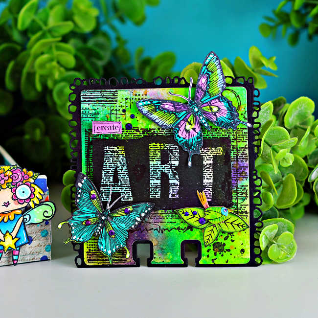 Colorful mixed media display featuring a vibrant AALL & Create MemoryDex card on the right and a decorated storage box on the left. The MemoryDex card has a neon green and purple Distress Oxide spray background with bold black “ART” lettering, hand-colored butterflies in teal and purple, and a small green bird with purple rhinestone accents. Beside it, a whimsical teal box is filled with bright florals and features a red-haired mermaid illustration, playful characters, and layered collage elements. The scene is styled with lush green foliage against a teal backdrop.