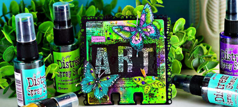 Mixed media AALL & Create MemoryDex card featuring a vibrant background created with Distress Oxide sprays in bright greens and purples. The card showcases bold “ART” lettering over layered text textures, accented with hand-colored butterflies shaded with Copic markers in teal, purple, and lime tones. The card sits among coordinating Distress Spray Stains and lush greenery, highlighting the bold, artistic color palette.