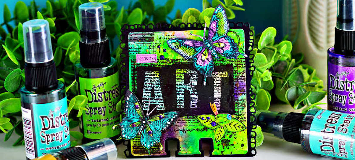 Mixed media AALL & Create MemoryDex card featuring a vibrant background created with Distress Oxide sprays in bright greens and purples. The card showcases bold “ART” lettering over layered text textures, accented with hand-colored butterflies shaded with Copic markers in teal, purple, and lime tones. The card sits among coordinating Distress Spray Stains and lush greenery, highlighting the bold, artistic color palette.