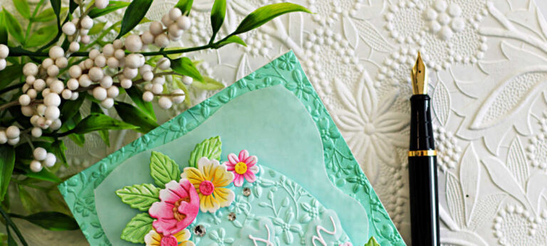 Elegant spring egg card featuring the Simon Says Stamp Country Basket Bouquet die set layered over Nested Eggs Dies and a Ribbon Loop Frame. The background and egg are dry embossed with the Tiny Flora Embossing Folder and softly ink blended in mint green. Pink and yellow ink-blended florals with leafy accents frame the egg, and a white “He is Risen” Script die sentiment sits centered on the embossed egg panel.
