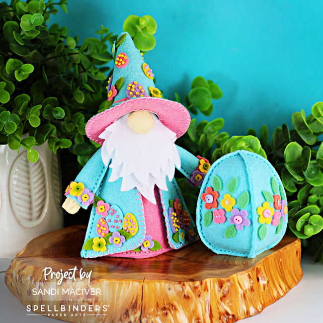Easter Fergus the Gnome handmade felt decor dressed in pastel teal and pink, featuring beaded felt eggs and floral embellishments on his coat and hat, displayed beside a matching 3D felt Easter egg with flowers on a wooden slice surrounded by greenery against a turquoise background.