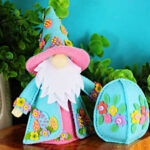 Handmade Easter Fergus the Gnome felt decor featuring a teal cone hat and matching coat decorated with beaded felt eggs and colorful felt flowers, a white beard, and pink accents, styled on a wooden slice with greenery against a turquoise background