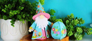 Handmade Easter Fergus the Gnome felt decor featuring a teal cone hat and matching coat decorated with beaded felt eggs and colorful felt flowers, a white beard, and pink accents, styled on a wooden slice with greenery against a turquoise background