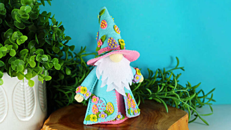Handmade Easter Fergus the Gnome felt decor featuring a teal cone hat and matching coat decorated with beaded felt eggs and colorful felt flowers, a white beard, and pink accents, styled on a wooden slice with greenery against a turquoise background.
