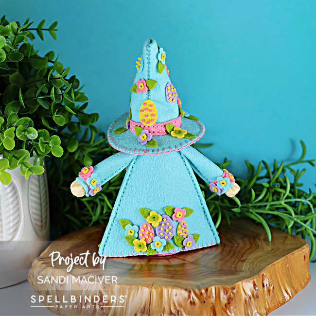 Back view of handmade Easter Fergus the Gnome felt decor showing a teal felt coat and cone hat embellished with beaded felt eggs and layered felt flowers, pink stitched trim, and floral details along the hem, styled on a wooden slice with greenery against a turquoise background.