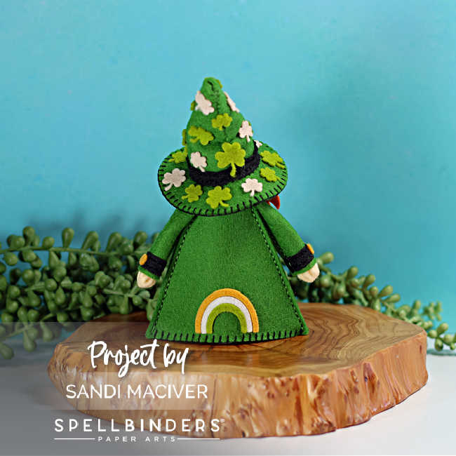 Back view of handmade St. Patrick’s Day Fergus the Gnome felt decor featuring a green felt coat and cone hat decorated with felt shamrocks and four-leaf clovers, black cuff details, and a small rainbow accent on the back of the coat, styled on a wooden slice with greenery against a teal background.