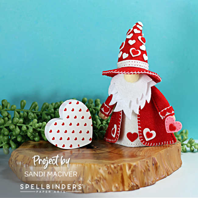 Handmade Valentine’s Day Fergus the Gnome felt decor wearing a red cone hat and matching coat decorated with white and pink hearts, featuring a white felt beard and heart accents, styled beside a layered felt heart on a wooden slice with greenery against a teal background.