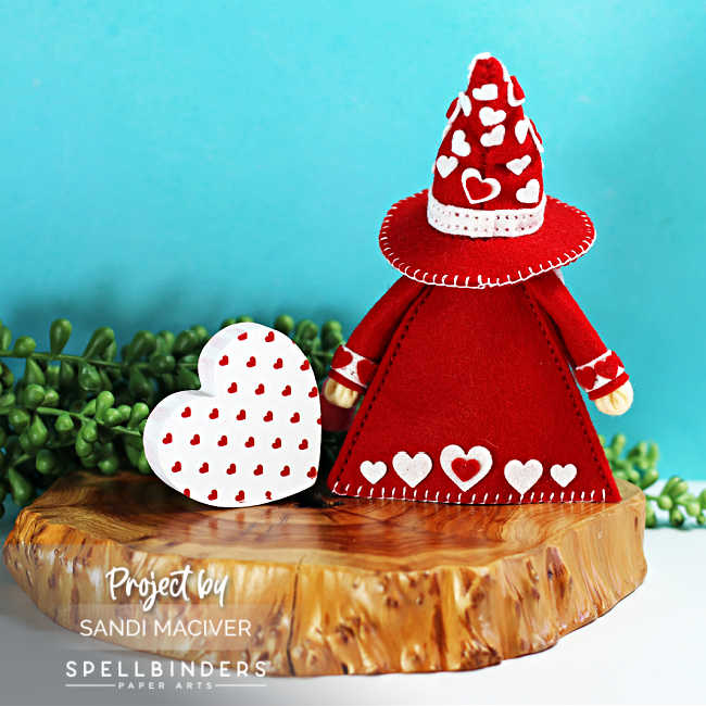 Back view of handmade Valentine’s Day Fergus the Gnome felt decor showing a red felt coat and cone hat embellished with white and pink heart details, stitched trim, and heart accents along the hem, styled beside a layered felt heart on a wooden slice with greenery against a teal background.