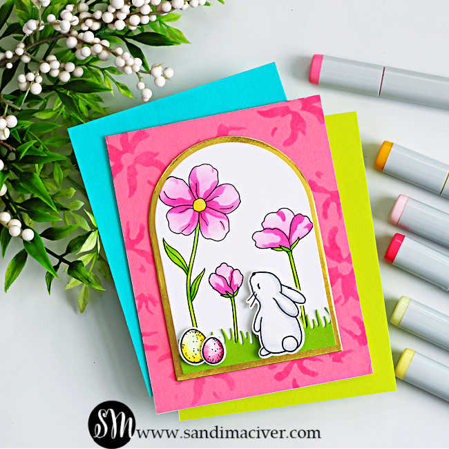Handmade spring card featuring a white bunny standing in green grass and looking up at pink flowers inside a gold-trimmed arched window. Two pastel Easter eggs sit at the bunny’s feet. The scene is layered over a pink stenciled background with teal and lime cardstock mats, photographed on a white surface with alcohol markers and greenery accents