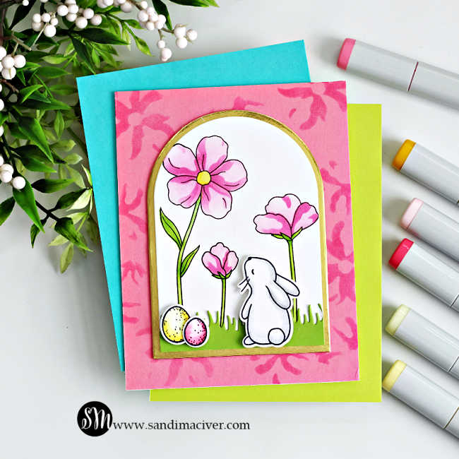 Handmade spring card featuring a white bunny standing in green grass and looking up at pink flowers inside a gold-trimmed arched window. Two pastel Easter eggs sit at the bunny’s feet. The scene is layered over a pink stenciled background with teal and lime cardstock mats, photographed on a white surface with alcohol markers and greenery accents.