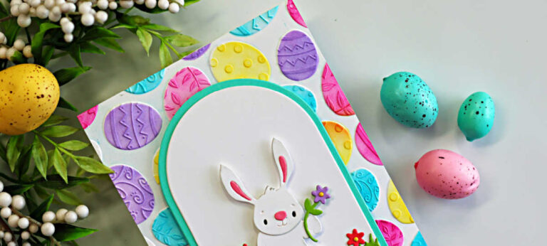 Handmade Easter card featuring a white die-cut bunny holding a purple flower, standing in grass with bright red and purple flowers inside a teal arched frame. The background is covered in colorful embossed Easter eggs in pink, yellow, teal, and purple. Styled with spring greenery and decorative pastel eggs around the card.