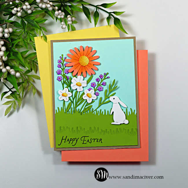 Handmade Easter card featuring layered die-cut spring flowers and a white bunny in a grassy garden scene. The card has a soft aqua sky background, bright coral and white flowers with green foliage, and a “Happy Easter” sentiment stamped along the bottom, created using the My Monthly Hero February 2026 kit.