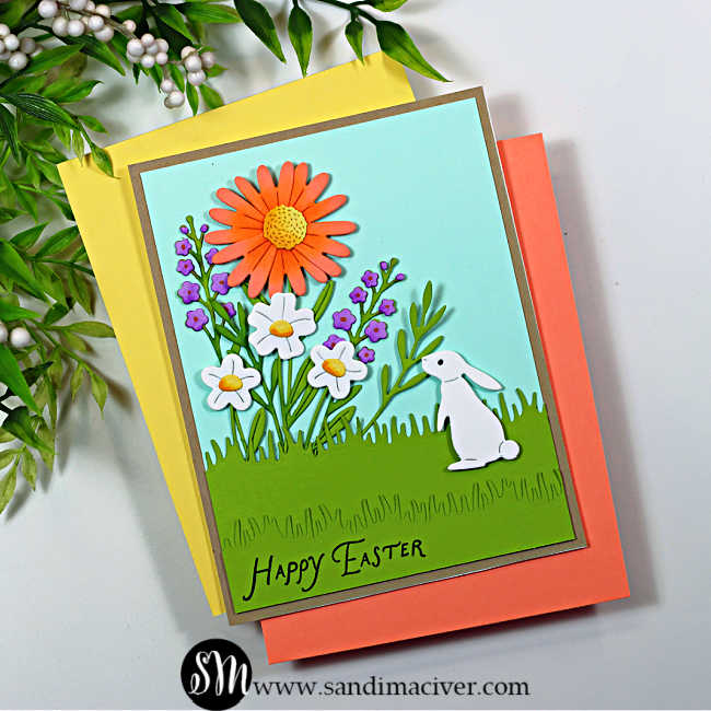 Handmade Easter card featuring layered die-cut spring flowers and a white bunny in a grassy garden scene. The card has a soft aqua sky background, bright coral and white flowers with green foliage, and a “Happy Easter” sentiment stamped along the bottom, created using the My Monthly Hero February 2026 kit.