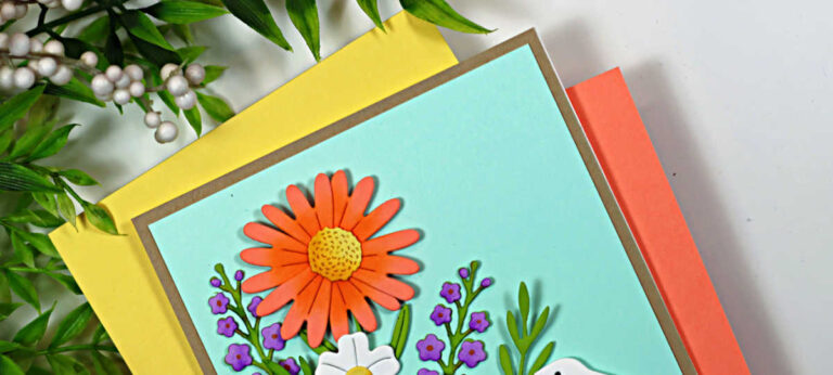 Handmade Easter card featuring layered die-cut spring flowers and a white bunny in a grassy garden scene. The card has a soft aqua sky background, bright coral and white flowers with green foliage, and a “Happy Easter” sentiment stamped along the bottom, created using the My Monthly Hero February 2026 kit.