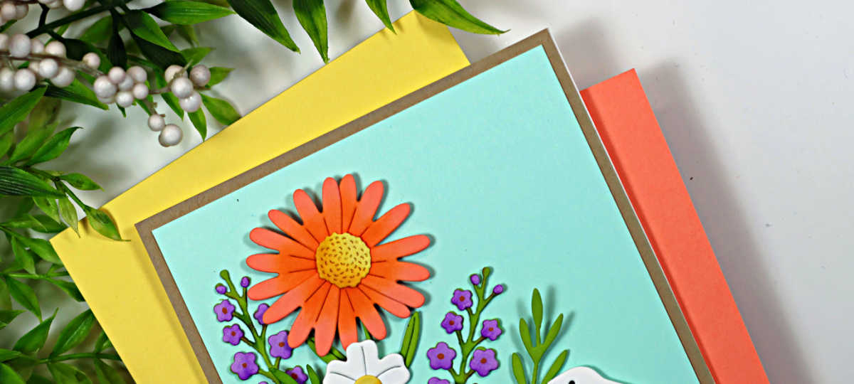 Handmade Easter card featuring layered die-cut spring flowers and a white bunny in a grassy garden scene. The card has a soft aqua sky background, bright coral and white flowers with green foliage, and a “Happy Easter” sentiment stamped along the bottom, created using the My Monthly Hero February 2026 kit.