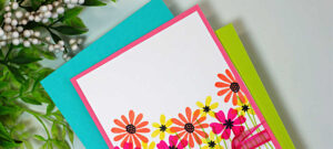 One-layer handmade thank-you card featuring colorful stamped wildflowers from the My Monthly Hero Wildflower Stamp and Cut set. Pink, coral, and yellow flowers with black centers rise from simple green stems on a white background, accented with a bright pink ribbon bow across the lower third and a stamped “Thanks a Bunch” sentiment.