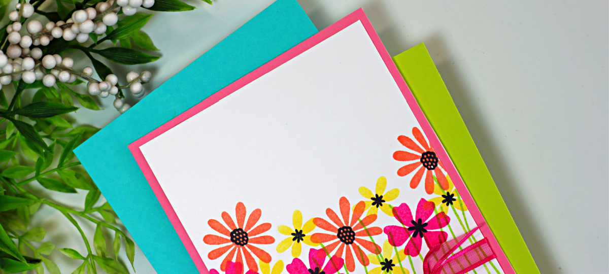 One-layer handmade thank-you card featuring colorful stamped wildflowers from the My Monthly Hero Wildflower Stamp and Cut set. Pink, coral, and yellow flowers with black centers rise from simple green stems on a white background, accented with a bright pink ribbon bow across the lower third and a stamped “Thanks a Bunch” sentiment.
