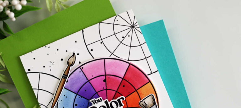 A handmade A2 card featuring the Pop of Color stamp and die set from Simon Says Stamp. The design showcases a vibrant rainbow color wheel ink blended in bright hues of pink, purple, blue, green, yellow, and orange. The sentiment “You Color My World” is layered in the center of the wheel. Three realistic paint brushes with brown handles are arranged around the circle, all popped up with dimension. The white background is stamped with overlapping black color wheel outlines and splattered with black ink for artistic detail. The card is layered over bright green and teal cardstock panels.