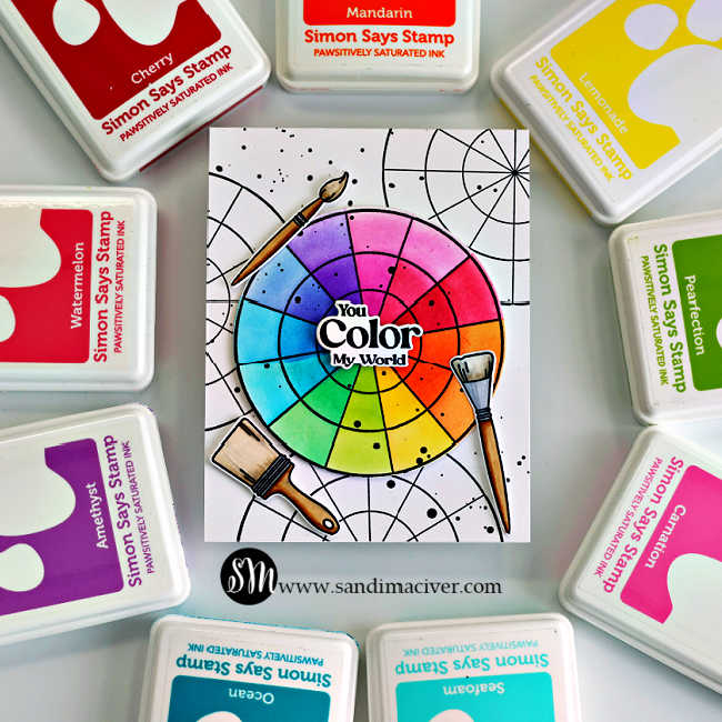 A handmade A2 card featuring a bright rainbow color wheel created with the Simon Says Stamp Pop of Color stamp and die set. The ink-blended wheel includes vibrant shades of pink, purple, blue, green, yellow, and orange with the layered sentiment “You Color My World” in the center. Three realistic paint brushes with brown handles and silver ferrules are arranged around the wheel. The white background is stamped with overlapping black color wheel outlines and black ink splatter for artistic detail. The card is photographed flat lay style and surrounded by Simon Says Stamp Pawsitively Saturated Ink pads in coordinating colors including Cherry, Mandarin, Lemonade, Pearfection, Carnation, Amethyst, Ocean, and Seafom.