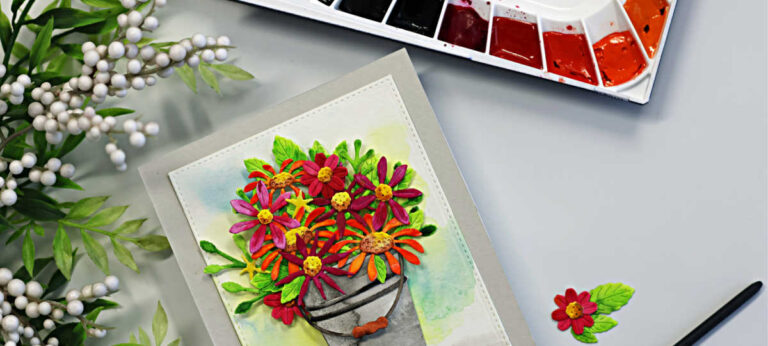 Handmade floral card featuring the Simon Says Stamp Watercolor Wildflower Bucket die set, with colorful die-cut wildflowers watercolored in bright pinks, oranges, yellows and greens arranged in a gray metal bucket. The background is softly watercolor blended in blue and green tones, with a small sentiment strip reading “wishing you joy always.” A paintbrush and watercolor palette are styled beside the card.