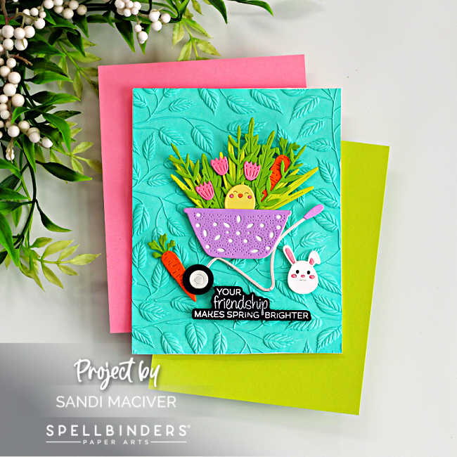 Handmade Easter card featuring a teal embossed leafy background layered over pink and lime green cardstock. A purple pierced wheelbarrow filled with die-cut greenery, pink tulips, orange carrots, and a yellow chick sits in the center. A small white bunny and carrot accent the design, along with the sentiment “Your friendship makes spring brighter.” Created with the Spellbinders February 2026 Small Die of the Month.