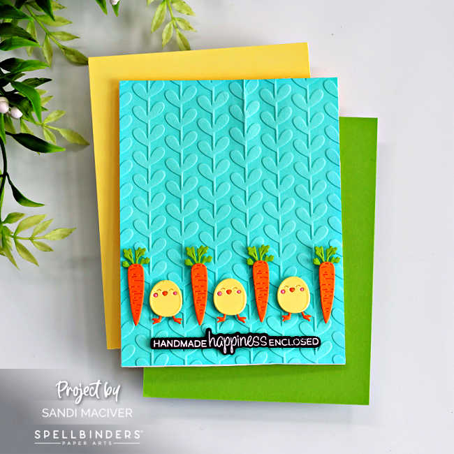 Handmade spring card featuring a teal embossed vine background layered over yellow and green cardstock. Along the bottom is a row of alternating die-cut orange carrots with green tops and round yellow chicks with tiny orange feet and pink cheeks. A black sentiment strip reads “Handmade happiness enclosed.” Created with the Spellbinders February 2026 Small Die of the Month.