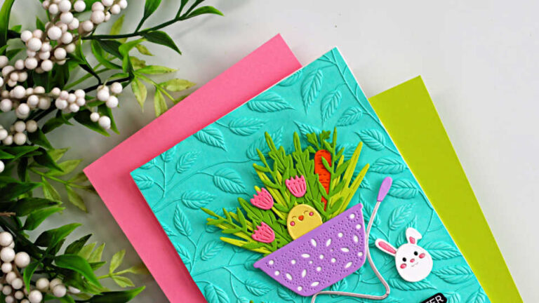 Close-up of a handmade Easter card created with the Spellbinders Small Die of the Month (February release). The card features a teal embossed leafy background layered over bright pink and lime green cardstock. A purple pierced basket filled with layered die-cut greenery, pink tulips, and orange carrots sits in the center, with a cute yellow chick peeking out. A small white bunny embellishment and decorative greenery accents complete the cheerful spring design.