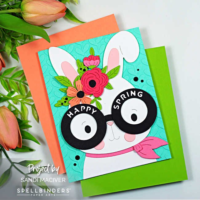 Square handmade spring card featuring a white bunny with oversized round glasses that read “Happy Spring,” layered die-cut flowers between the ears, and a pink scarf. The bunny is set against a turquoise embossed background, with coral and green cardstock mats peeking out underneath.