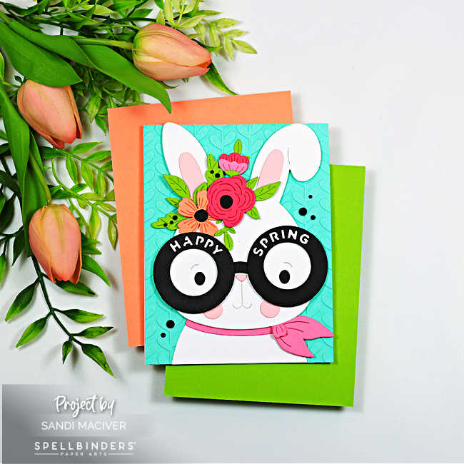 Square handmade spring card featuring a white bunny with oversized round glasses that read “Happy Spring,” layered die-cut flowers between the ears, and a pink scarf. The bunny is set against a turquoise embossed background, with coral and green cardstock mats peeking out underneath.