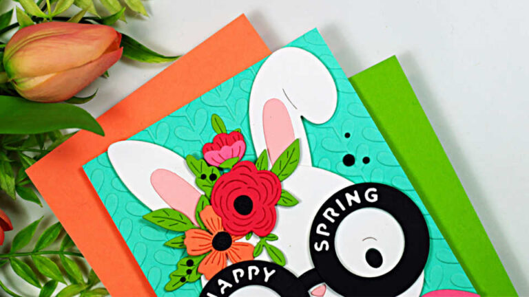 Square handmade spring card featuring a white bunny with oversized round glasses that read “Happy Spring,” layered die-cut flowers between the ears, and a pink scarf. The bunny is set against a turquoise embossed background, with coral and green cardstock mats peeking out underneath.