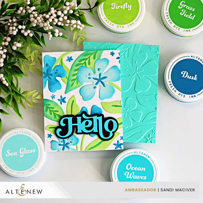 A handmade A2 card created using the Altenew Joyful Garden stencil and embossing folder set. The card features layered blue and teal hibiscus-style flowers with bright green leaves, stenciled over an embossed white background for added texture and dimension. A bold, layered “Hello” sentiment in black and teal is centered across the floral design. The card is photographed flat lay style with green and teal cardstock layers beneath it.