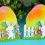 Free-standing Easter egg-shaped card with rainbow ink blending, die cut bunny, flowers, and white picket fence on a teal background