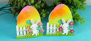 Free-standing Easter egg-shaped card with rainbow ink blending, die cut bunny, flowers, and white picket fence on a teal background