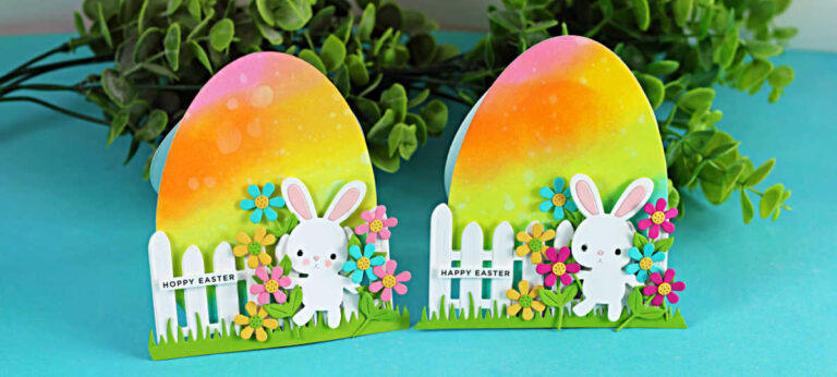 Free-standing Easter egg-shaped card with rainbow ink blending, die cut bunny, flowers, and white picket fence on a teal background