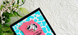 A handmade birthday card created with the Hero Arts My Monthly Hero March 2026 kit. The card features a black A2 card base layered with a bright turquoise cow-print background. Centered on the card is a dimensional pink milk carton die cut, with a cute black-and-white cow overlay attached to the front. The sentiment “It’s Your Birthday” is stamped in black near the bottom of the carton. The card is photographed on a white embossed floral background with greenery and white berries in the upper corner.