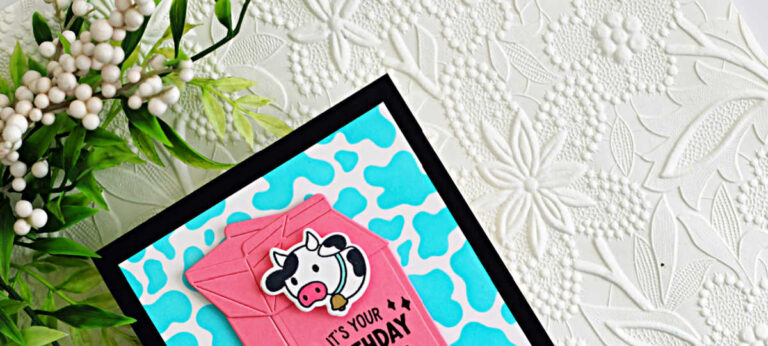 A handmade birthday card created with the Hero Arts My Monthly Hero March 2026 kit. The card features a black A2 card base layered with a bright turquoise cow-print background. Centered on the card is a dimensional pink milk carton die cut, with a cute black-and-white cow overlay attached to the front. The sentiment “It’s Your Birthday” is stamped in black near the bottom of the carton. The card is photographed on a white embossed floral background with greenery and white berries in the upper corner.